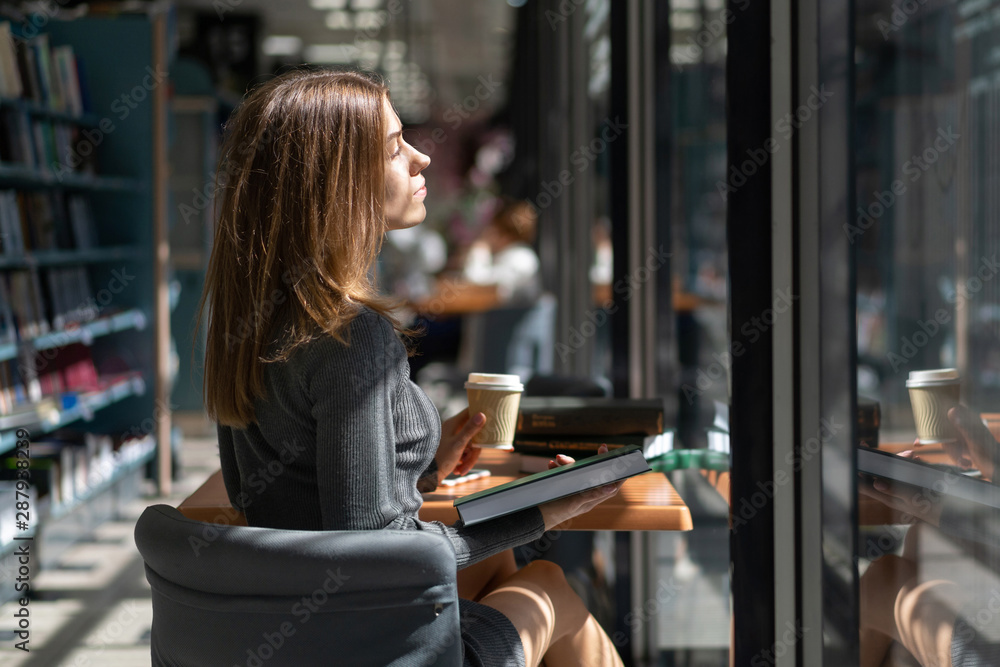 Fototapeta premium Caucasian girl student reads book in institute library and drinks coffee