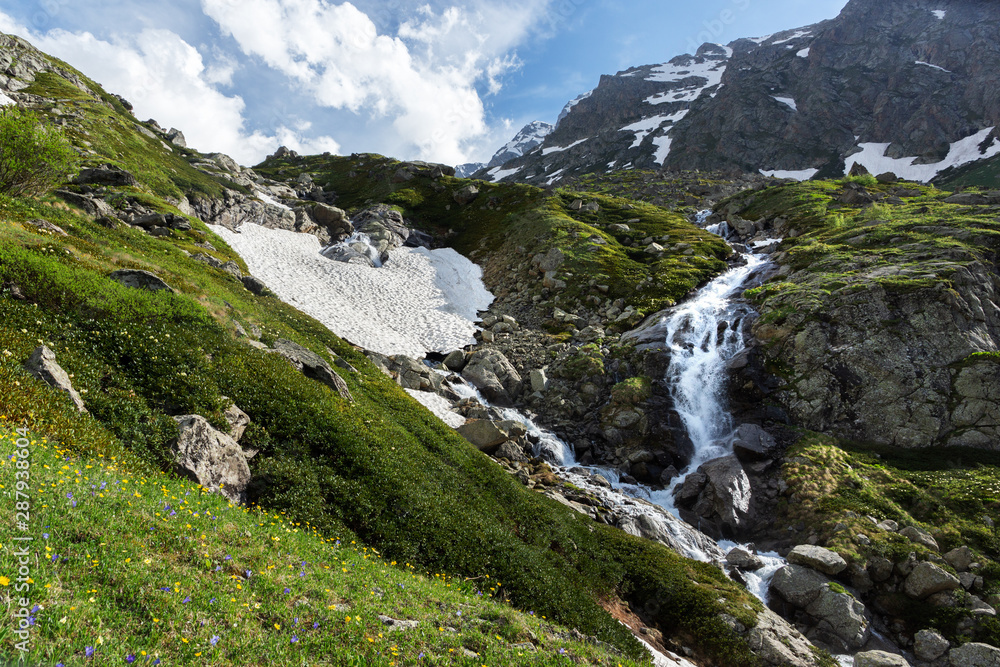 waterfall and glacier in the mountains in summer, flowering meadow, contrast between snow and green grass waterfall and glacier in the mountains in summer, flowering meadow, contrast between snow and green grass