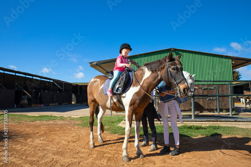 little girl learning to ride a horse outside of stable in a equestrian school 