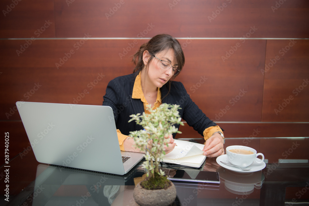 Attractive young businesswoman working in the office with laptop, cell phone and notebook and with a cup of coffee, with elegant wooden background Attractive young businesswoman working in the office with laptop, cell phone and notebook and with a cup of coffee, with elegant wooden background