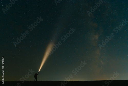Konstfotografi Back view of man with flashlight standing on green grassy field under beautiful dark blue summer starry sky