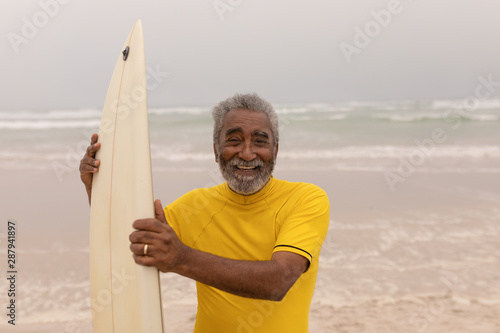 Senior male surfer standing with surfboard and looking at camera on the beach 