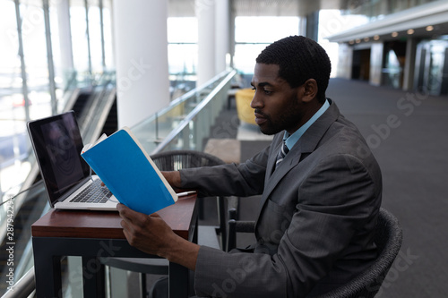 Side view of African-American businessman with laptop sitting at table and reading a book in modern 