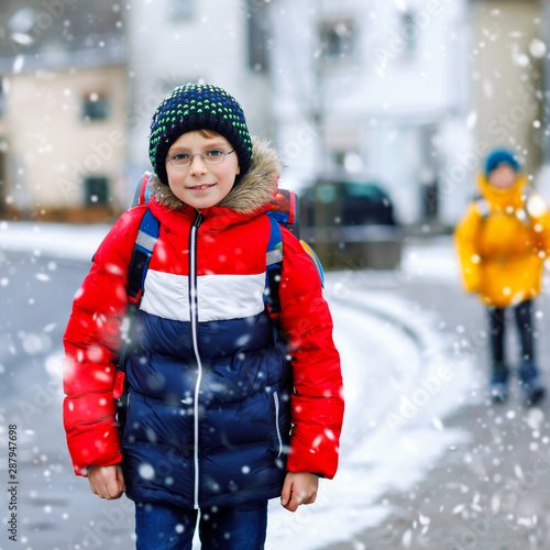 Little school kid boy of elementary class walking to school during snowfall. Happy child and student with eye glasses backpack in colorful winter clothes. schoolkid in yellow jacket on background