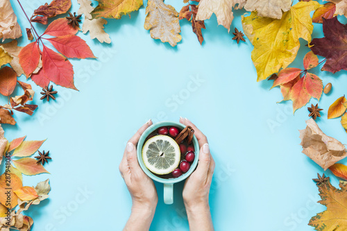 Cozy cup of healthy drink with cranberry in female hand and colorful fall leaves around on blue. Autumn composition. Fall pattern. Top view.