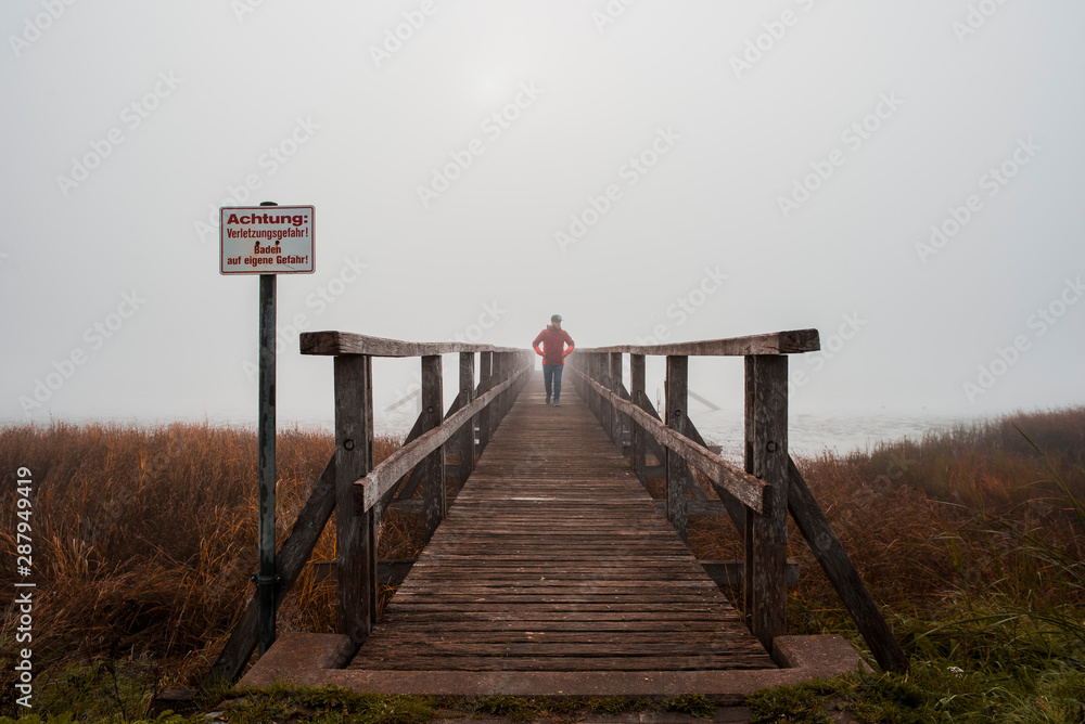 Fototapeta premium Steg im Nebel mit Mann in roter Jacke und Cap