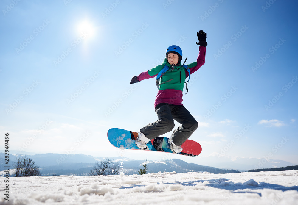 Young female snowboarder jumping in air on copy space background of clear bright blue sky and snowy mountains on sunny winter day. Winter sports and recreation, leisure outdoor activities concept.