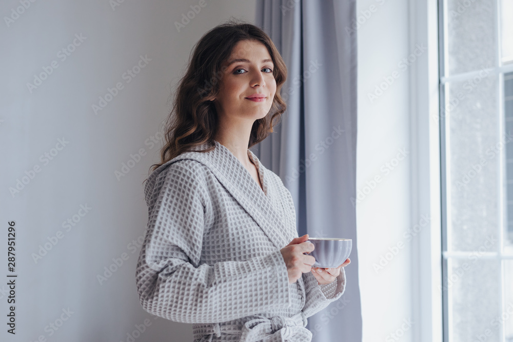 Portrait of pretty woman in bathrobe enjoying morning standing by the window and holding cup of tea. Portrait of pretty woman in bathrobe enjoying morning standing by the window and holding cup of tea.