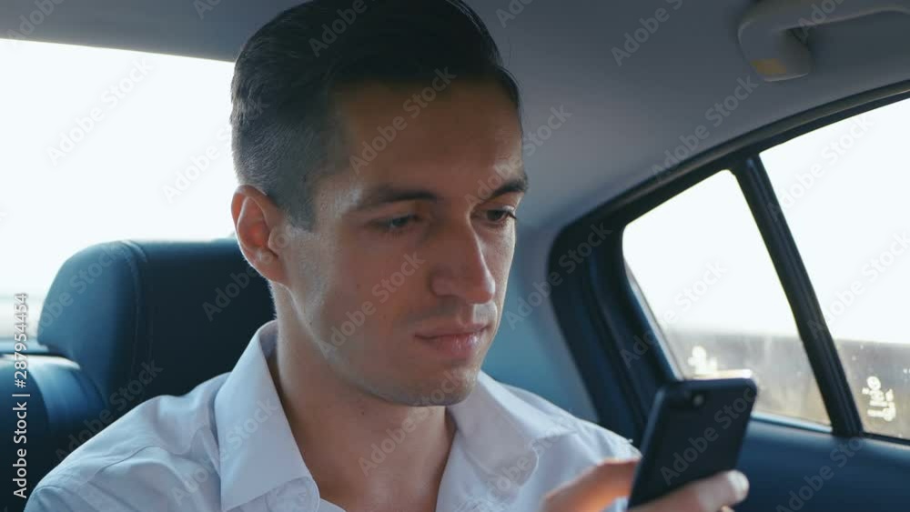 Young Businessman using his Smartphone during Traveling in a Car with a Driver. Man is checking Mails, Chats or the New Online while he rides in a Taxi Young Businessman using his Smartphone during Traveling in a Car with a Driver. Man is checking Mails, Chats or the New Online while he rides in a Taxi