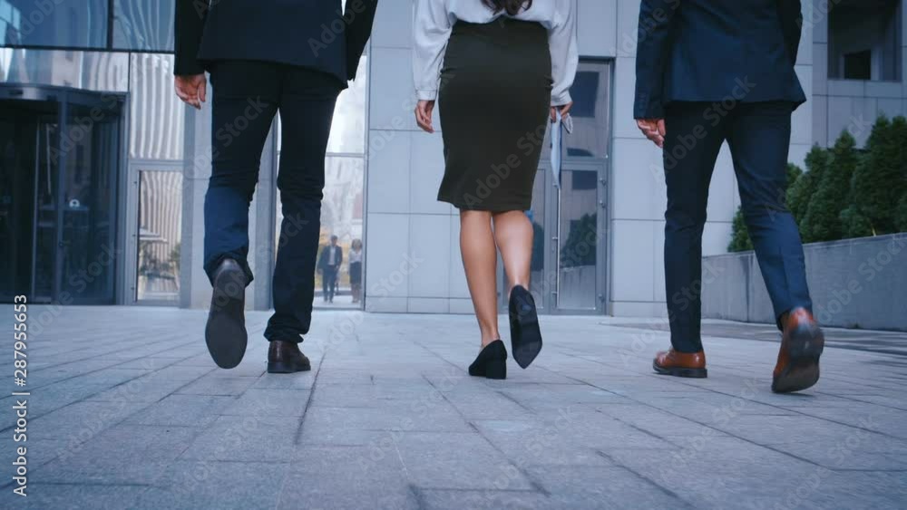 Rear back view feet of three successful business people commuting to work to office building. Businessmen and businesswoman in suits going in the city outdoor.