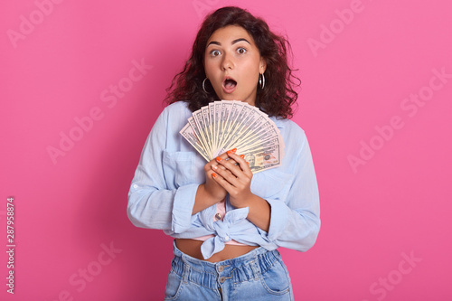 Close up portrait of attractive woman holding fan of money in her hands, keeps mouth opened, wearing stylish clothng, wins lottery, has shocked facial expression, wide open eyes with surprise. photo