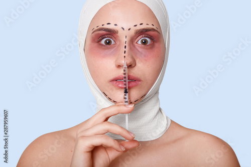 Shot of shocked scrared woman staring with bugged eyes, ready for recieves injection, afraids of plastic surgery, has marked lines on problematic zones of face, isolated over blue studio background photo