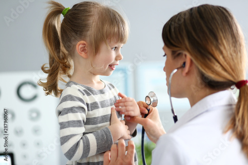 Portrait of cheerful child covering chest by hands. Smiling baby in striped sweater visiting family doctor. Health care and prevention concept. Blurred background