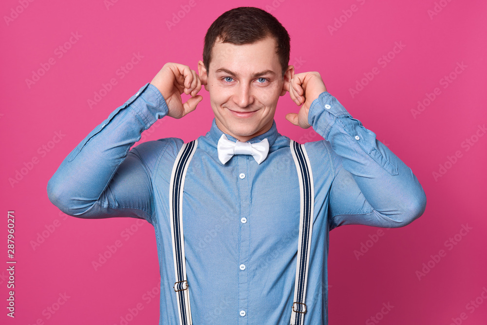 Portrait of handsome young man with charming smile, covering ears with fingers, does not want to hear his girlfriend, posing isolated on pink background in studio. People and relationship concept. Portrait of handsome young man with charming smile, covering ears with fingers, does not want to hear his girlfriend, posing isolated on pink background in studio. People and relationship concept.