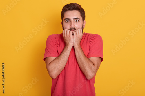 Studio shot of young man wearing red t shirt standing isolated over yellow background, guy looking scared, having amazed expression with hands under chin, bites his finger, sees something terrible.