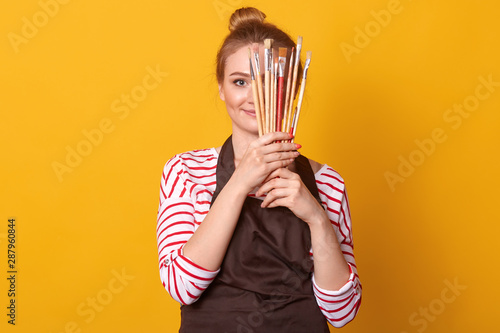 Horizontal shot of girl artist holds brushes in hands and hides behind it, lady dresses stripes casual shirt and brown apron, blonde woman with bunch, posing isolated over yellow studio background. photo