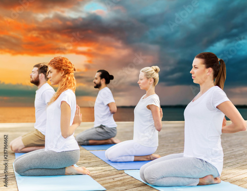 yoga, fitness, sport, and healthy lifestyle concept - group of people making hero pose on mat outdoors on sea pier over sunset background
