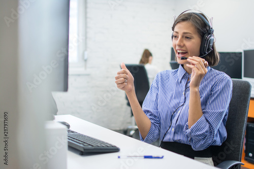 Beautiful friendly female costumer service operator showing thumb up to computer screen during video call in call centre