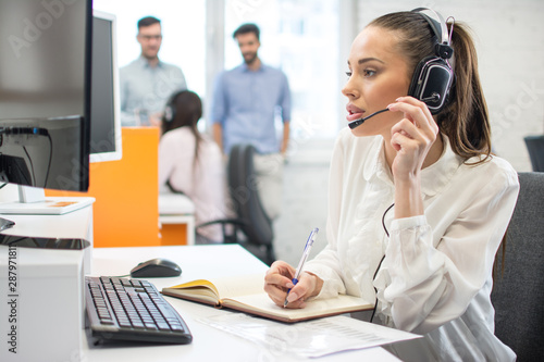 Beautiful young customer support female operator talking with client via hands-free headphones with microphone and writing notes to notebook in call center