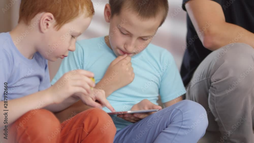 Handheld shot of male primary school teacher in glasses watching over cute schoolboys playing on tablet during recess Handheld shot of male primary school teacher in glasses watching over cute schoolboys playing on tablet during recess