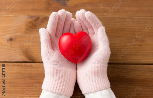 winter  valentine s day and christmas concept - hands in pale pink woollen gloves holding red heart over wooden boards background