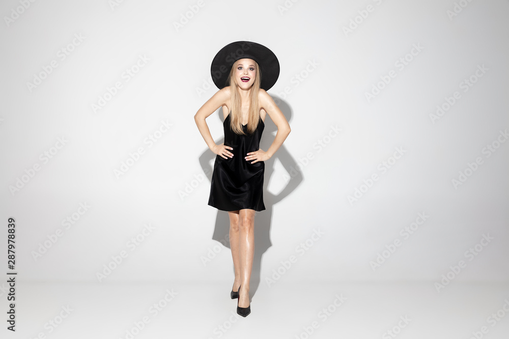 Young blonde woman in black hat and costume on white background. Attractive caucasian female model posing. Halloween, black friday, cyber monday, sales, autumn concept. Copyspace. Happy, astonished.