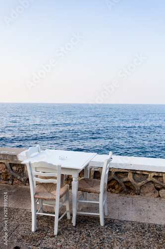 Weiße leere Stühle und Tisch aus Holz stehen an einer Strandpromenade mit Ausblick aufs Meer