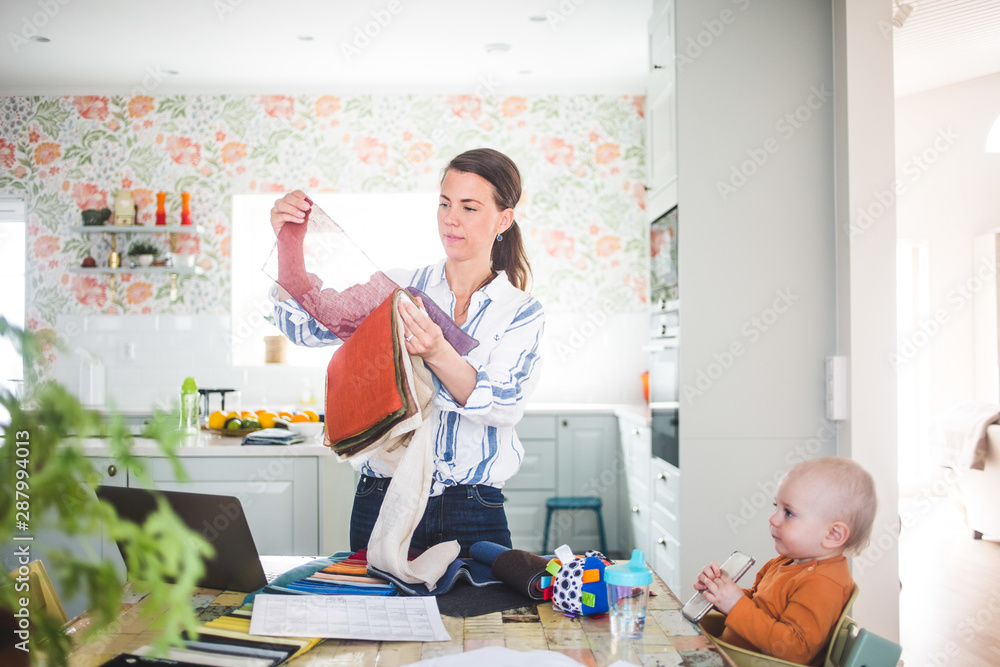Working mother holding fabric swatch while baby girl looking away in kitchen at home office Working mother holding fabric swatch while baby girl looking away in kitchen at home office
