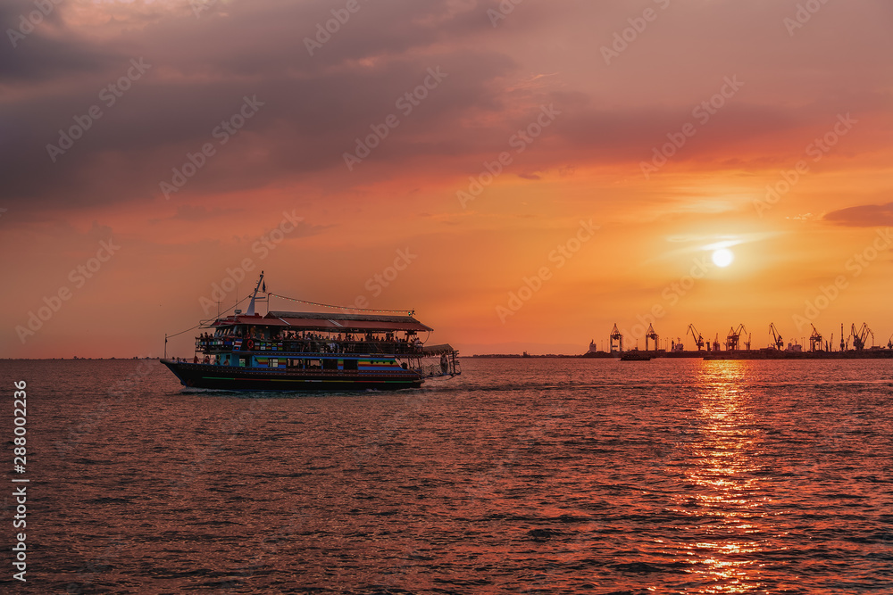 Tourist ship sailing during a golden hour sunset at Thessaloniki, Greece. Unidentified crowd aboard a small ship at Thermaikos gulf during orange sunset, with port cranes background.