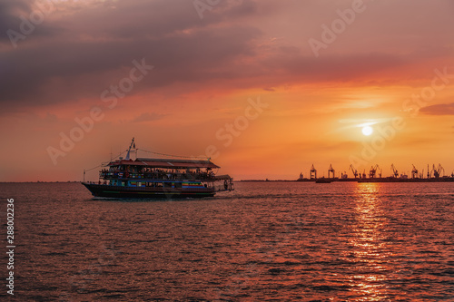 Tourist ship sailing during a golden hour sunset at Thessaloniki  Greece. Unidentified crowd aboard a small ship at Thermaikos gulf during orange sunset  with port cranes background.
