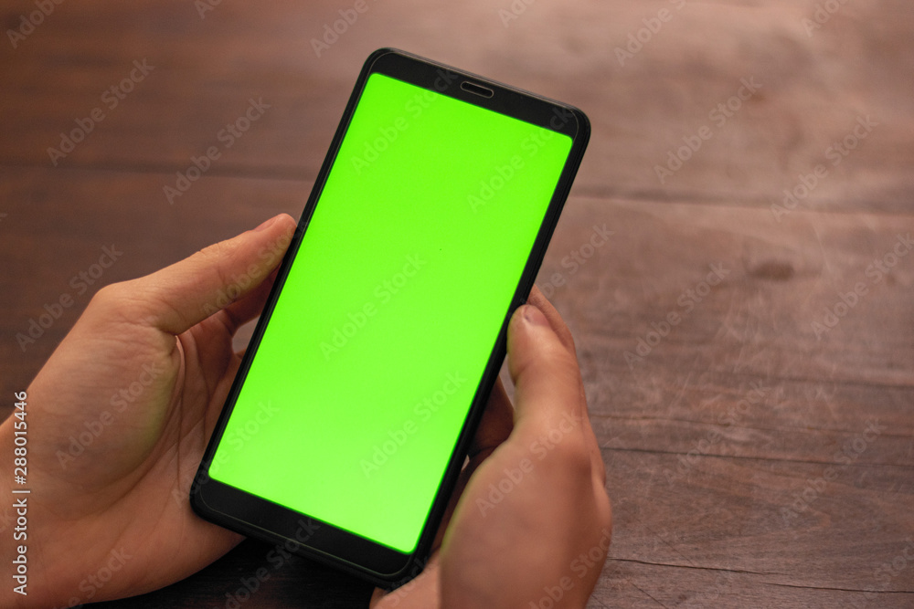 Hand of caucasian person holding a black unidentified smartphone with green screen on a wooden table background