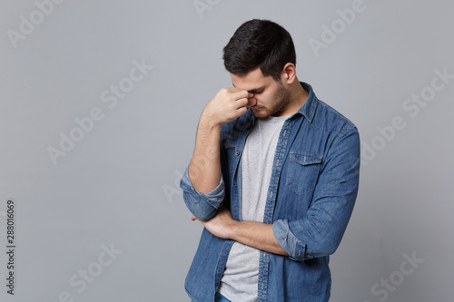 Handsome tired stylish unshaven young man in denim jeans shirt posing isolated on grey wall background studio portrait. People lifestyle concept. Mock up copy space. keep eyes closed, put hand on nose © ViDi Studio