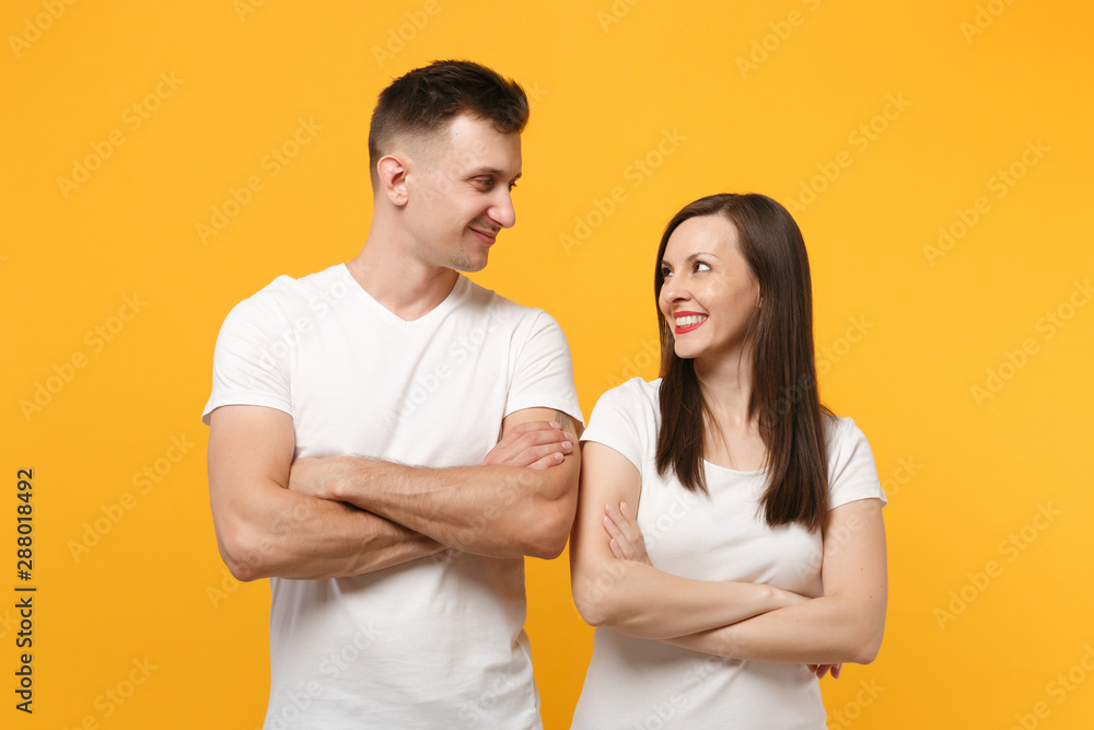 Young couple two friends guy girl in white empty blank t-shirts posing isolated on yellow orange background. People lifestyle concept. Mock up copy space. Holding hands crossed, looking at each other. Young couple two friends guy girl in white empty blank t-shirts posing isolated on yellow orange background. People lifestyle concept. Mock up copy space. Holding hands crossed, looking at each other.