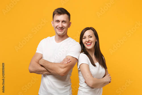 Smiling young couple two friends guy girls in white blank empty design t-shirts posing isolated on yellow orange wall background. People lifestyle concept. Mock up copy space. Holding hands crossed.