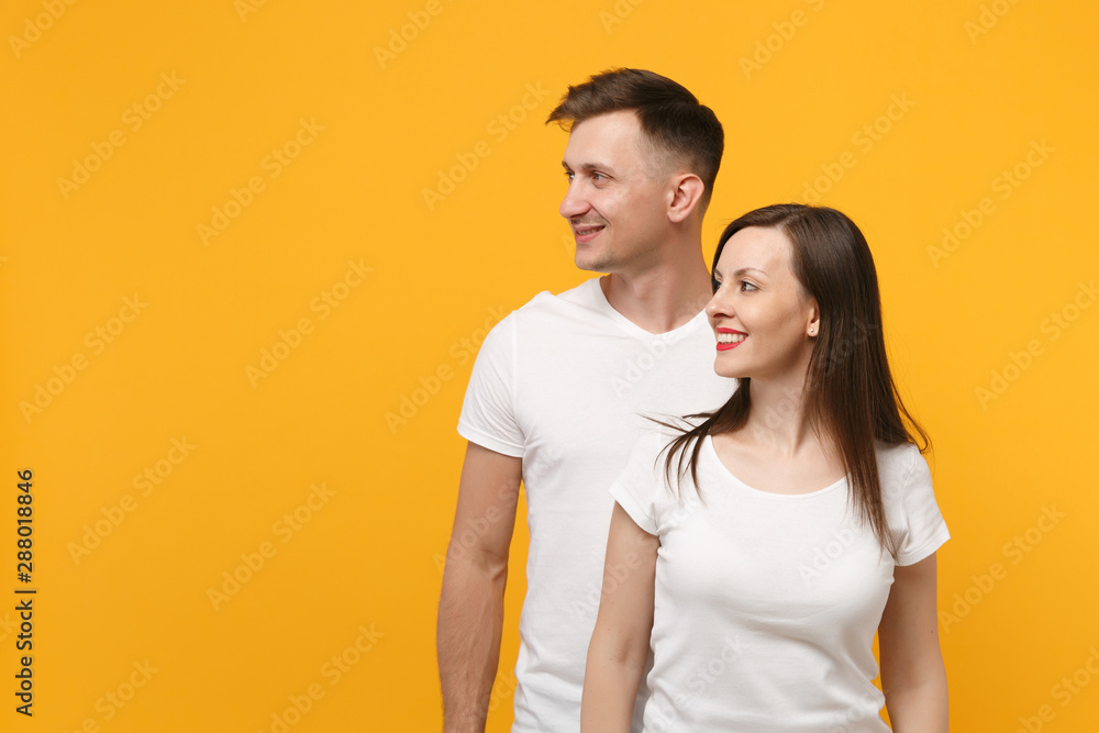 Beautiful smiling young couple two friends guy girl in white empty blank design t-shirts posing isolated on yellow orange wall background. People lifestyle concept. Mock up copy space. Looking aside. Beautiful smiling young couple two friends guy girl in white empty blank design t-shirts posing isolated on yellow orange wall background. People lifestyle concept. Mock up copy space. Looking aside.