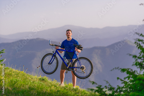 Fototapeta Naklejka Na Ścianę i Meble -  Tired cyclist is wiping his sweat off his face while pushing his bicycle uphill on a dirt road in a mountain.