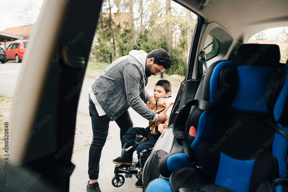 Father carrying disabled son from wheelchair seen from car Father carrying disabled son from wheelchair seen from car