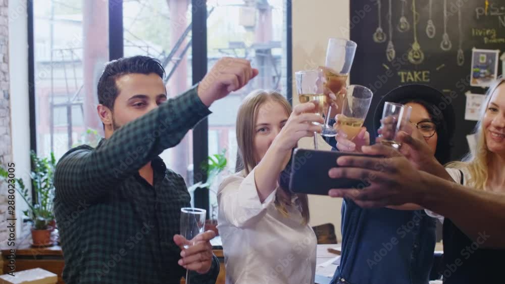 Joyous multiethnic team of young male and female designers smiling and toasting with champagne at smartphone camera while taking selfie together during party in the office Joyous multiethnic team of young male and female designers smiling and toasting with champagne at smartphone camera while taking selfie together during party in the office