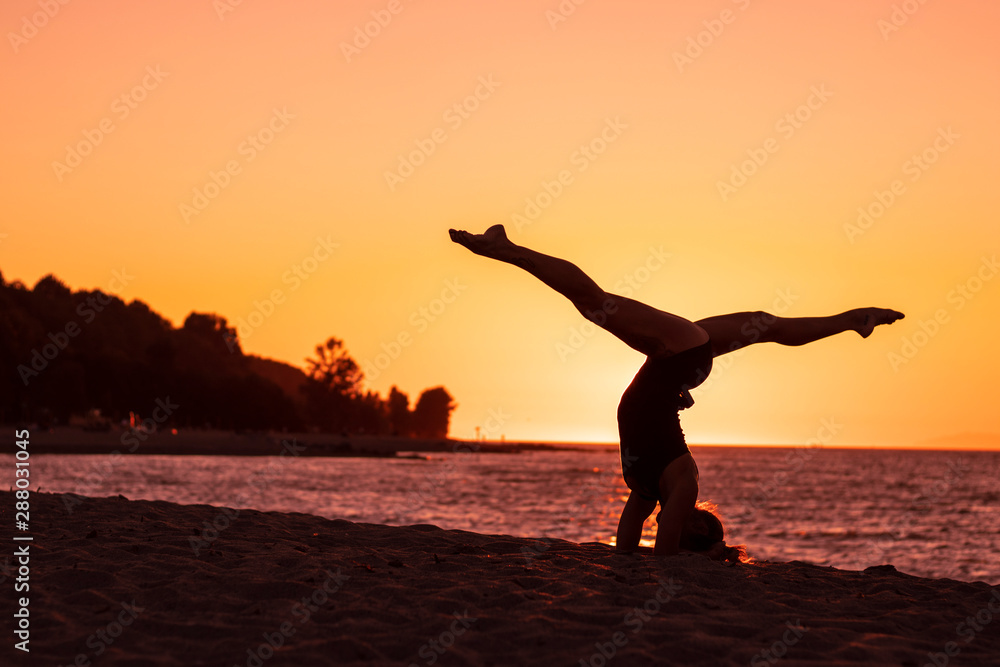 Flexible woman practicing yoga and gymnastics on the beach at sunset, in utthita pose, wellbeing, good posture, zen attitude, healthy, ocean pacific view and tree shadow backgrounds