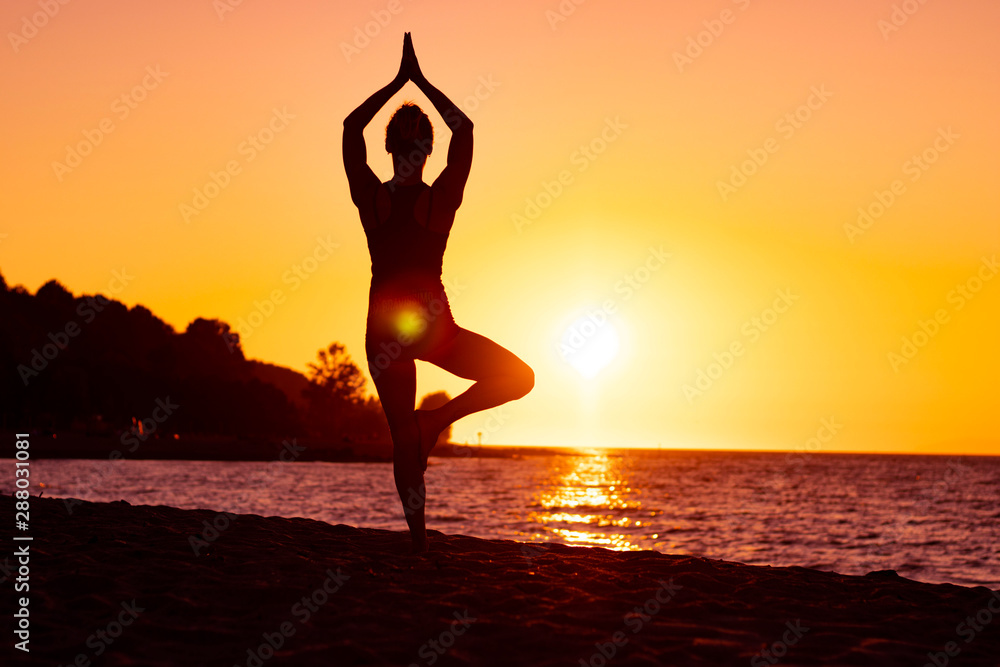 Flexible woman practicing yoga and gymnastics on the beach at sunset, in tree pose, well being, good posture, zen attitude, healthy, ocean pacific view