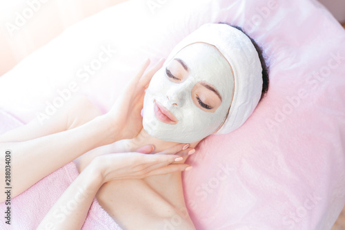 Portrait of beautiful woman laying with towel on the head. Young girl enjoys cream facial mask. Lady getting spa treatment at beauty salon. Cosmetology and skincare concept.