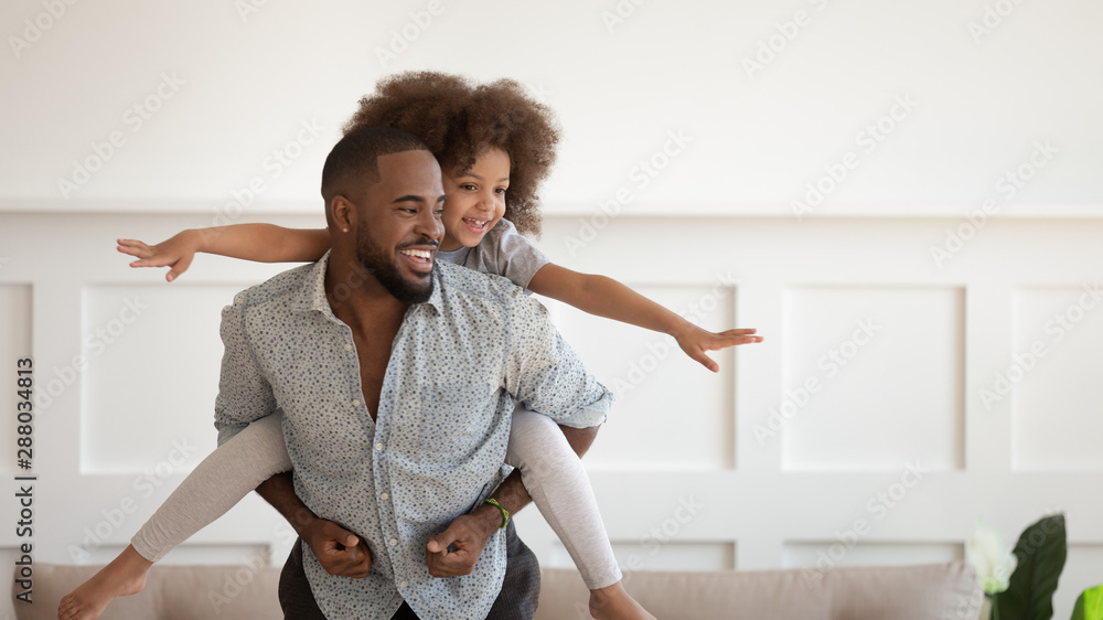 Excited black man giving piggyback ride to happy cute daughter. - obrazy, fototapety, plakaty 
