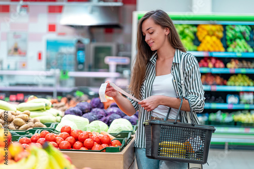 Foto Young woman with shopping basket checks and examines a sales receipt after purchasing food in a grocery store