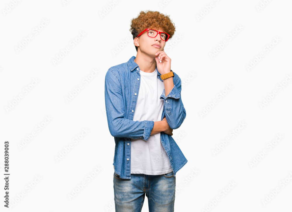 Young handsome man with afro hair wearing glasses with hand on chin thinking about question, pensive expression. Smiling with thoughtful face. Doubt concept. Young handsome man with afro hair wearing glasses with hand on chin thinking about question, pensive expression. Smiling with thoughtful face. Doubt concept.
