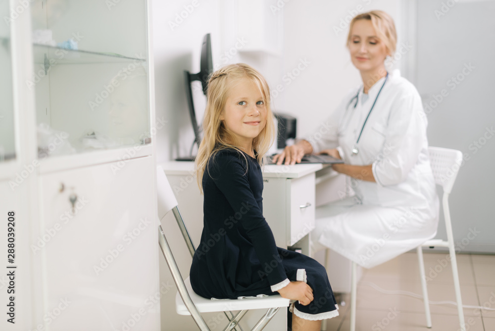 Fototapeta premium Cute kid girl with blond hair plays looking at the camera in pediatrician appointment. Doctor takes notes on a patient outpatient card.