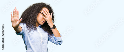 Young beautiful business girl with curly hair wearing glasses covering eyes with hands and doing stop gesture with sad and fear expression. Embarrassed and negative concept.