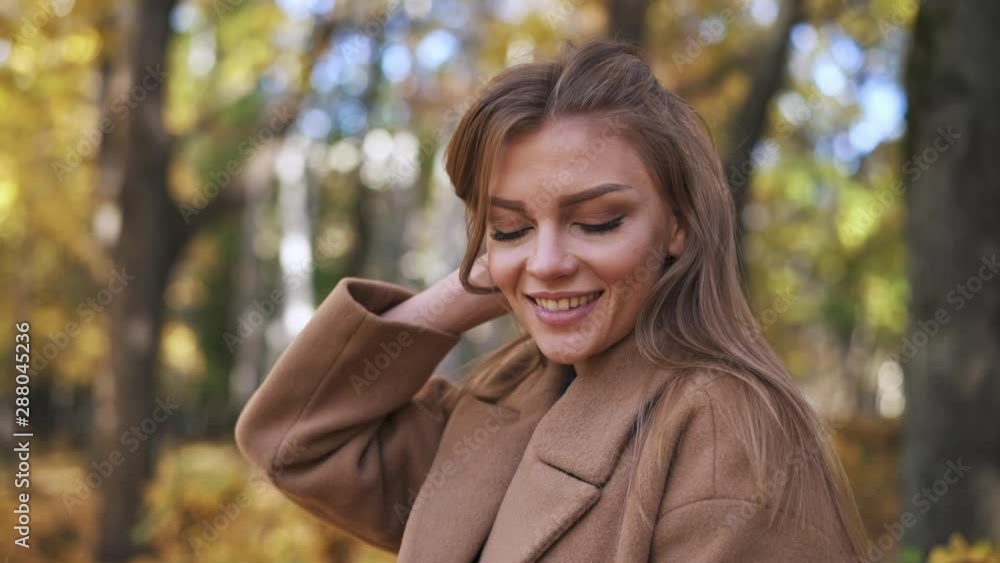 Smiling young woman with long blond hair playing with it in sunny autumn park. She is wearing beige coat. Positive emotions. Slider slow motion close up shot Smiling young woman with long blond hair playing with it in sunny autumn park. She is wearing beige coat. Positive emotions. Slider slow motion close up shot