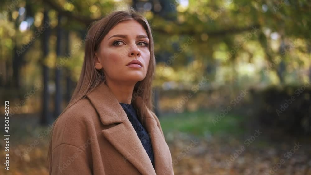 Worried and stressed young woman with long blond hair wearing beige coat looking around standing in autumn park. Slider slow motion medium shot. Worried and stressed young woman with long blond hair wearing beige coat looking around standing in autumn park. Slider slow motion medium shot.