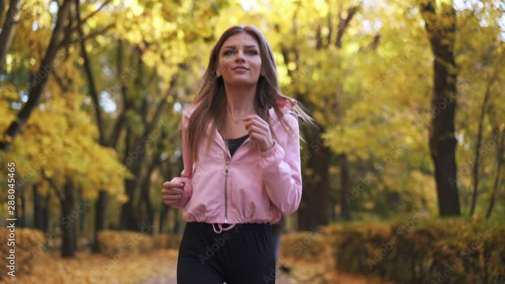 Happy young blonde woman in pink hoodie jogging in autumn city park. Concept of staying fit and healthy lifestyle. Tracking slow motion medium shot. Happy young blonde woman in pink hoodie jogging in autumn city park. Concept of staying fit and healthy lifestyle. Tracking slow motion medium shot.
