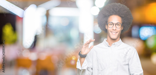 Young african american man with afro hair wearing glasses gesturing with hands showing big and large size sign, measure symbol. Smiling looking at the camera. Measuring concept.